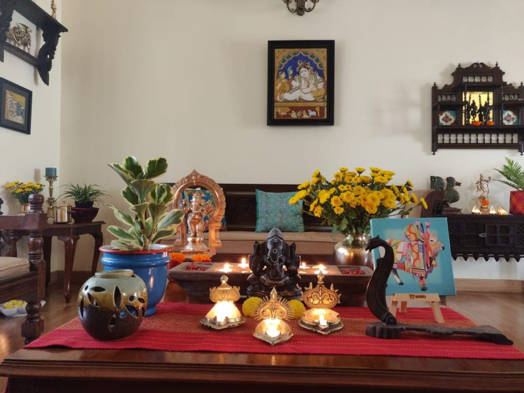 The table at the living room is setting up with fresh flowers, diyas, sculpture, green plants and Pichwai frame