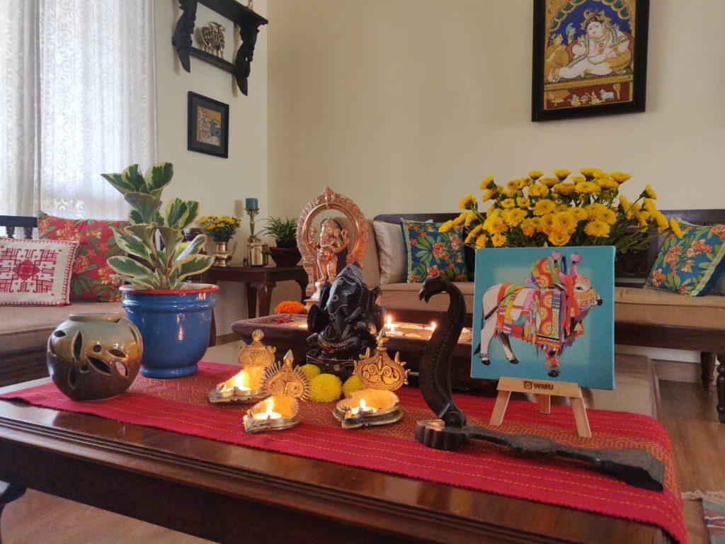 The table at the living room is setting up with fresh flowers, diyas, sculpture, green plants and Pichwai frame