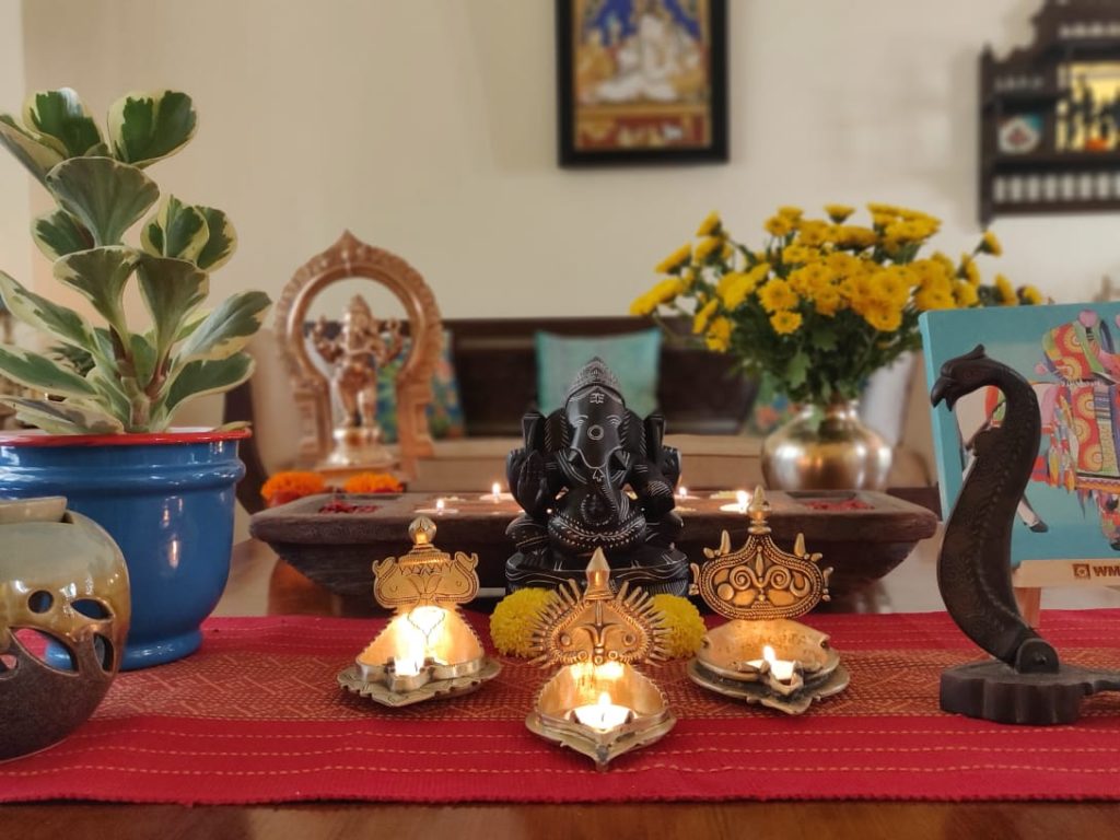 The table at the living room is setting up with fresh flowers, diyas, sculpture, green plants and Pichwai frame