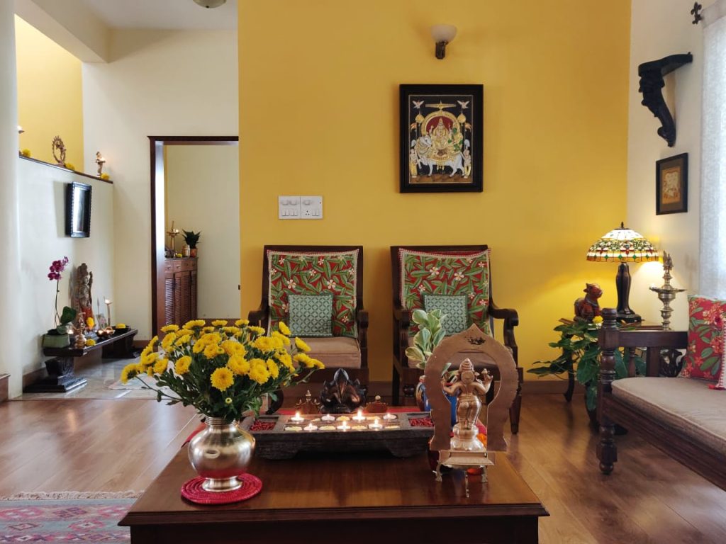 the living room is decorated with  brass, sculpture, green plants, fresh flowers and wooden diya stand and wall frames