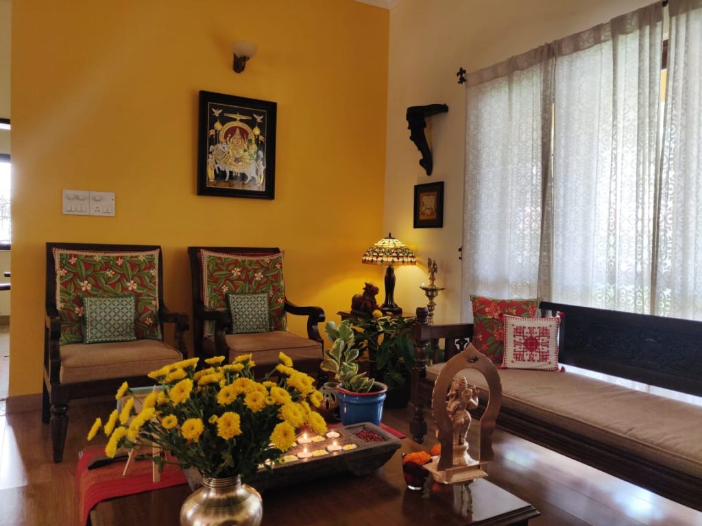 the living room is decorated with  brass, sculpture, green plants, fresh flowers and wooden diya stand and wall frames