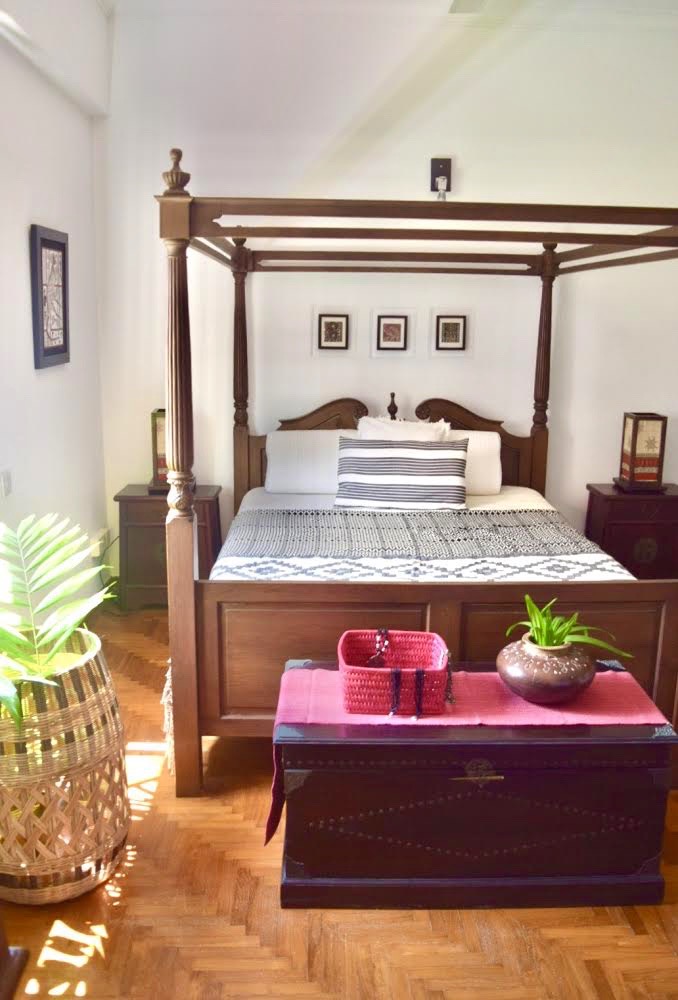 the master bedroom is decorated with four posters, wood trunk with red runner, basket and brass vase on it, green plants and bamboo table lamps at the corner of the room | ASHA RAJ home tour