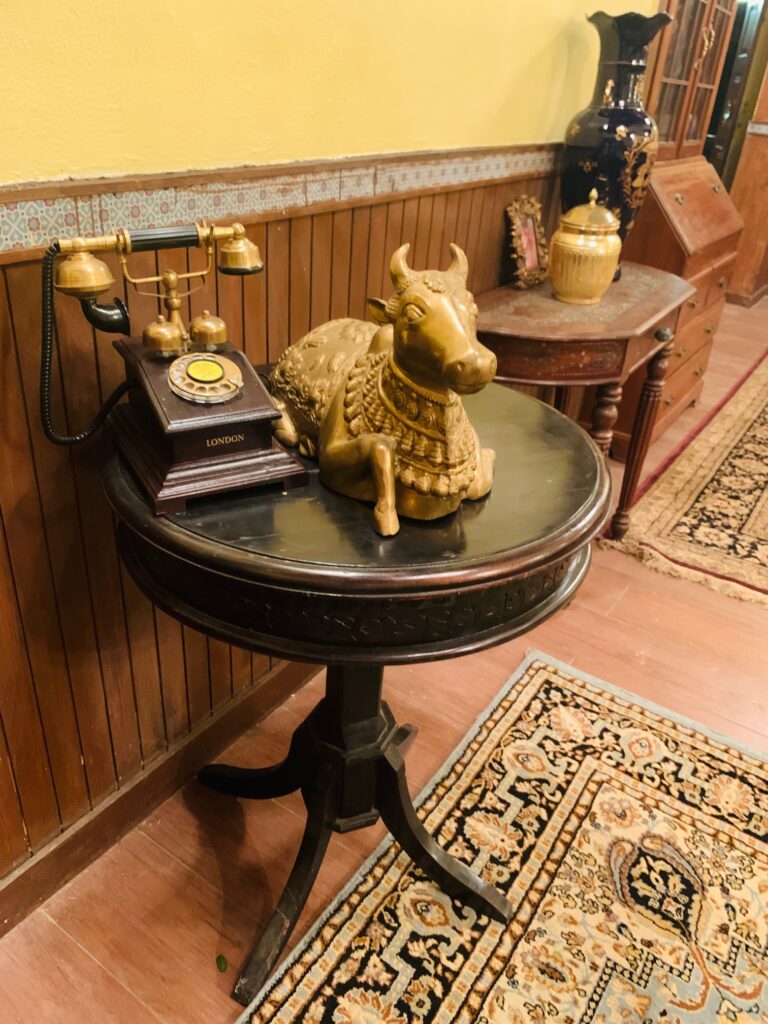Villa Rashmi - A Heritage Gem in Mumbai | Brass nandi statue, wooden brass antique telephone and brass jar box are on top of the table at the corner of the bedroom | TheKeybunch decor blog