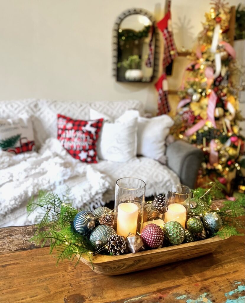 A Christmas centerpiece of a doughbowl filled with a candle, some plumosus fern greenery, pine cones and Christmas ball decor